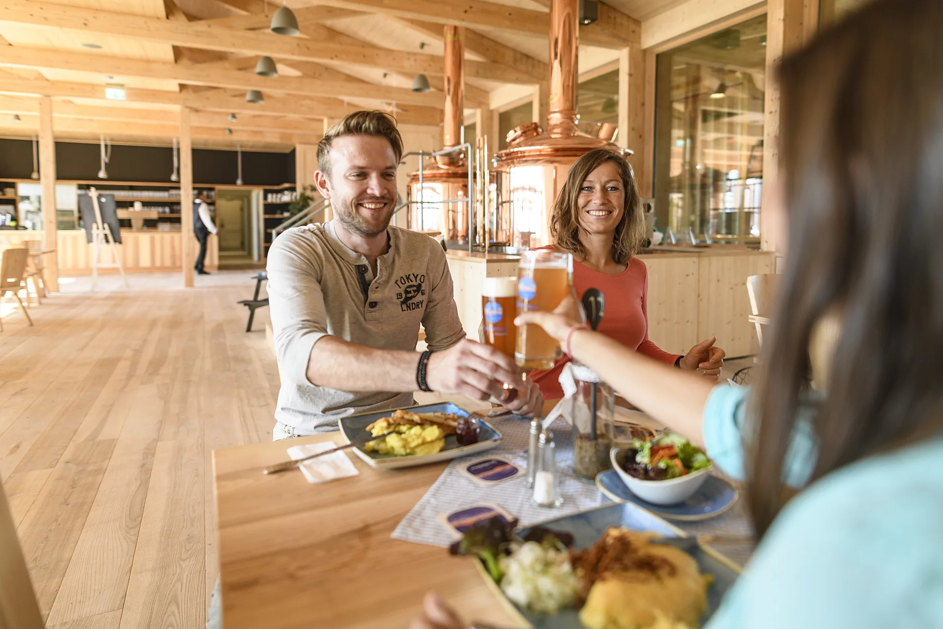 Gäste stoßen in der hellen Gaststube neben der kupfernen Schaubrauerei an